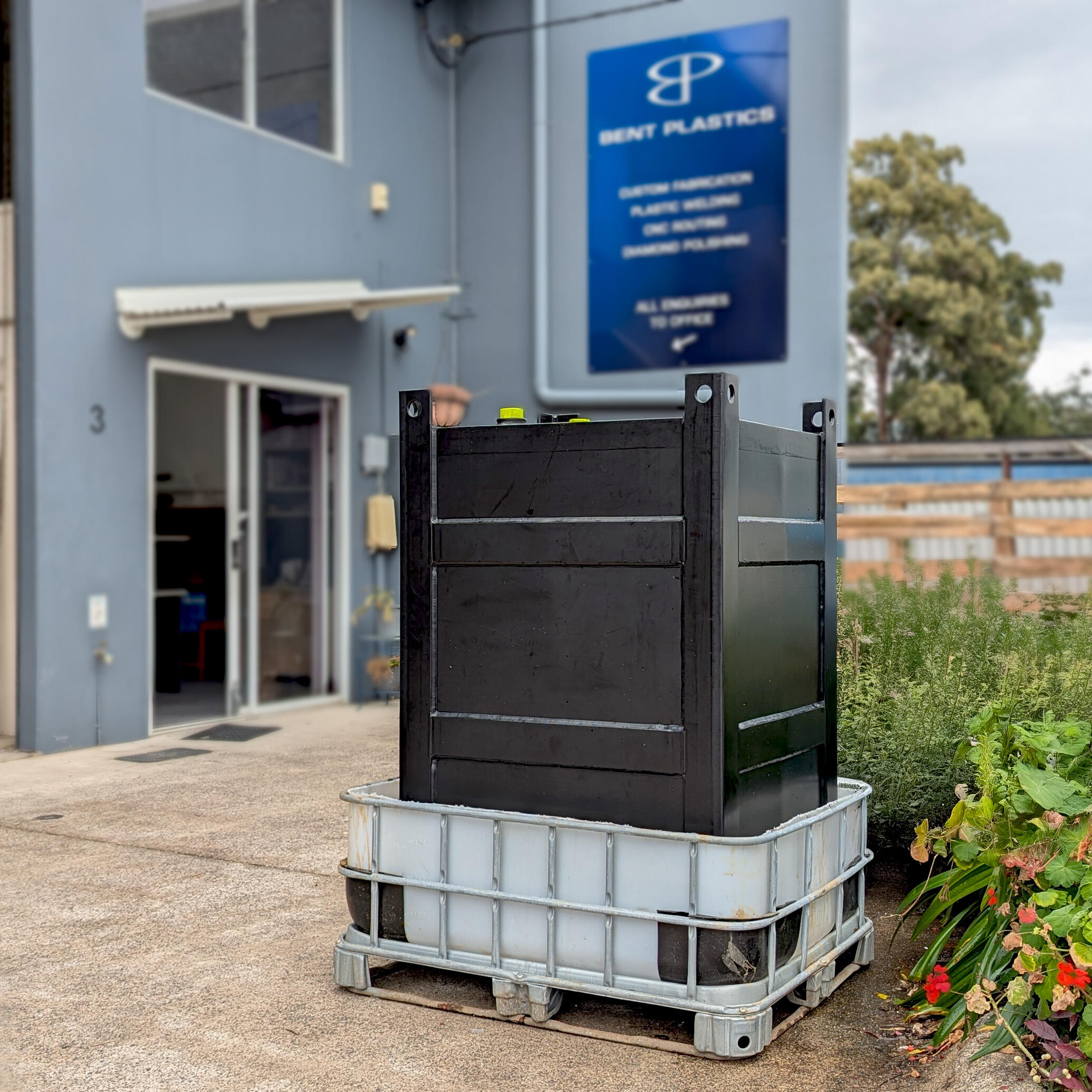 Bent Plastics Sunshine Coast Rectangular black plastic tank sitting in a metal and plastic tray, in front of a factory with a sign that says Bent Plastics